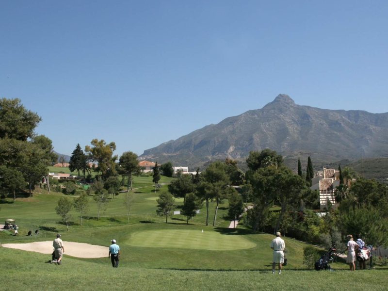 Golfers playing on a lush golf course in Marbella with panoramic views of La Concha mountain under a clear blue sky on the Costa del Sol.